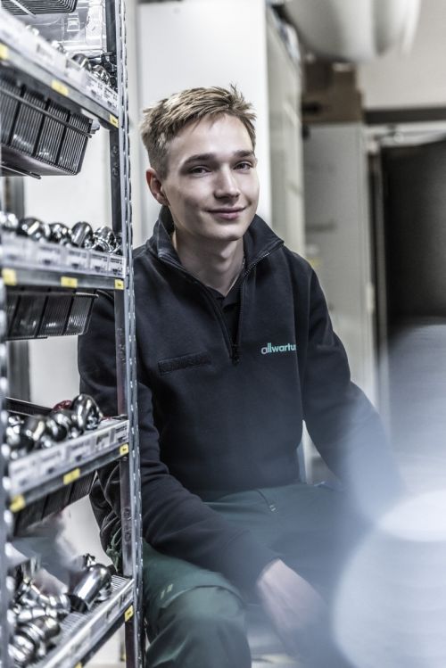 Ein junger Mann sitzt in einem Lager mit Sanitärteilen und trägt eine Jacke mit dem Schriftzug "allwartung".
