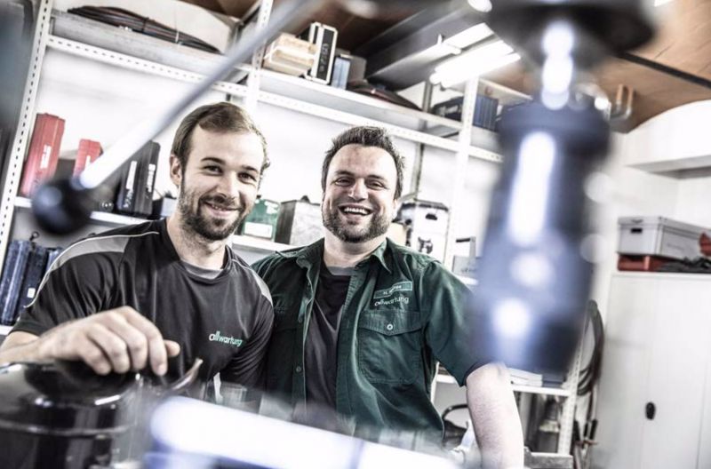Zwei lächelnde Handwerker stehen in einer Werkstatt, umgeben von Werkzeugen für Sanitär- und Klimatechnik.